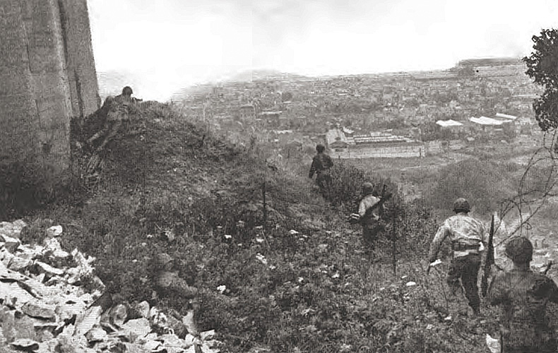 The main objective for U.S. troops in Normandy was the capture of Cherbourg with its commercial harbor. Here, infantrymen work their way around Fort du Roule to destroy seaward facing German artillery guns by using demolitions, thereby allowing American forces to sweep into the city below.