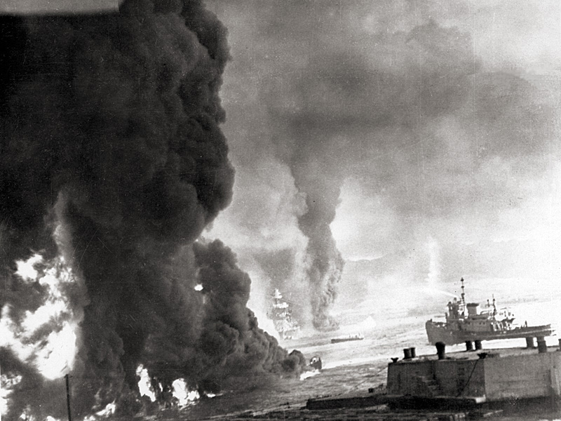 Oil fires burn on the water near Ford Island. USS Maryland is in the center background. A harbor tug is at right.