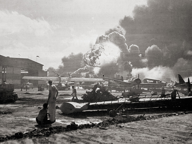 Sailors stand amid wrecked planes at the Ford Island seaplane base, watching as USS Shaw explodes.
