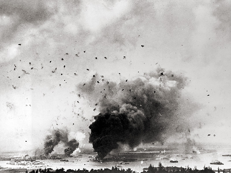 View of Pearl Harbor from the hills to the north shows anti-aircraft shells bursting overhead. Large column of smoke in lower center is from USS Arizona.