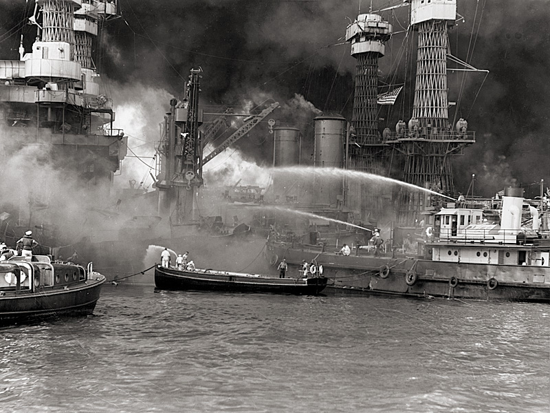 Fighting fires on the sunken battleship USS West Virginia. A garbage lighter is at right, with her crewmen aiming two fire hoses at the flames. Also assisting are a motor launch and an officer's motorboat.