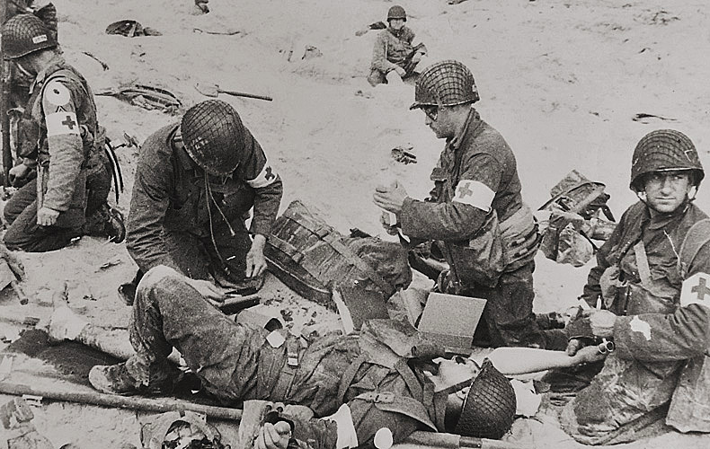 American medics render first aid to troops in the initial landing. In the background other troops dig into the soft sand of the beach.