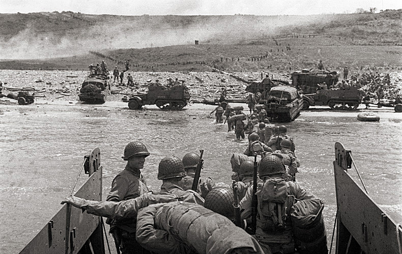 The steady flow of troops and equipment continues on Omaha Beach. Half-tracks are seen at the water line while lines of men are now headed inland.