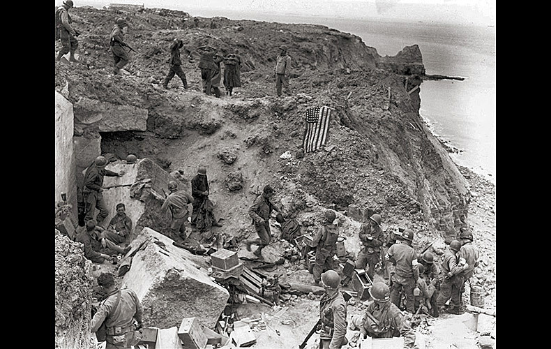 A view of Pointe du Hoc on June 8th, after relief forces reached the Army Rangers who secured the strategic cliffs on D-Day, thereby protecting American troops on Omaha Beach. The American flag was spread out to prevent friendly fire from American tanks. German prisoners are seen being moved after their capture by the relieving forces.